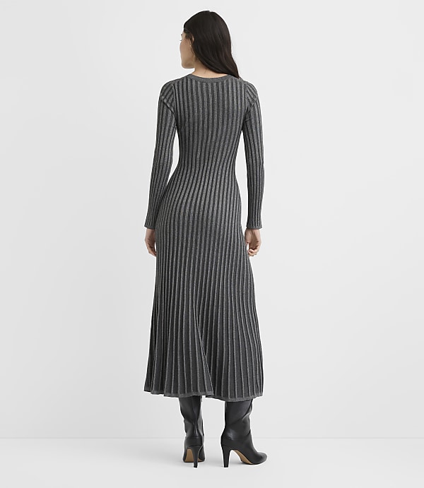 A woman wearing a long-sleeved, ribbed grey maxi dress and black heeled boots stands with her back to the camera against a plain white background.