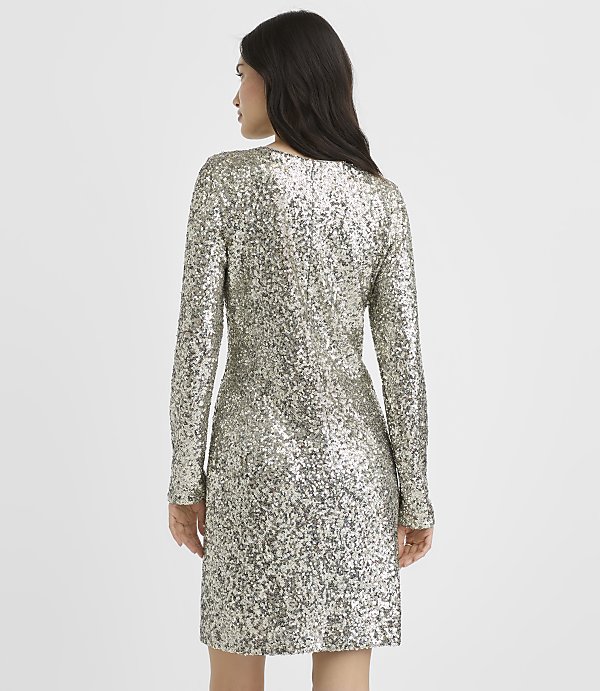 A woman wearing a long-sleeved silver sequin dress stands with her back to the camera against a plain white background.