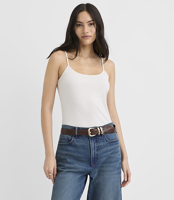 A woman wearing a white camisole and blue jeans with a brown belt stands against a plain white background.
