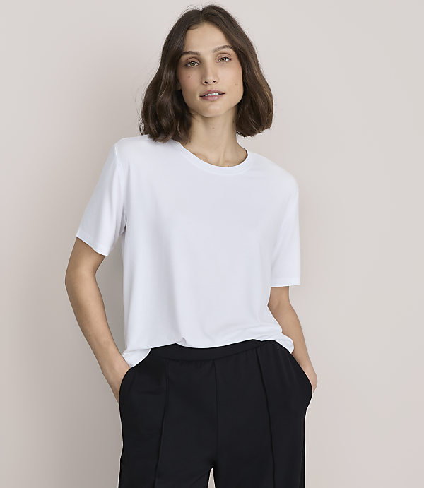 A woman wearing a white t-shirt and black trousers stands against a plain, light-beige background.