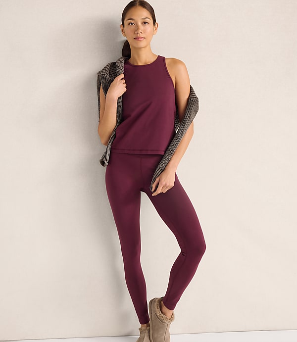 A woman wearing a maroon tank top and leggings with a patterned cardigan draped over her shoulders stands against a plain, light-colored wall.