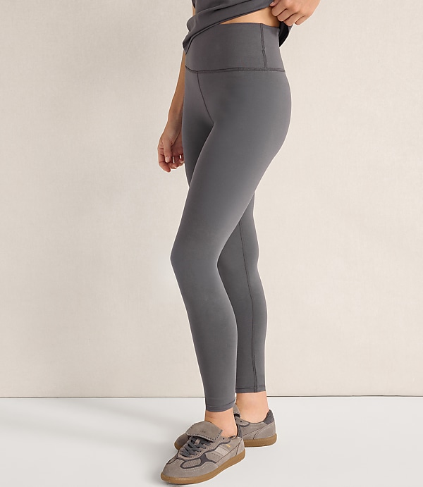 A person wearing grey leggings and sneakers stands against a plain, light-colored wall.