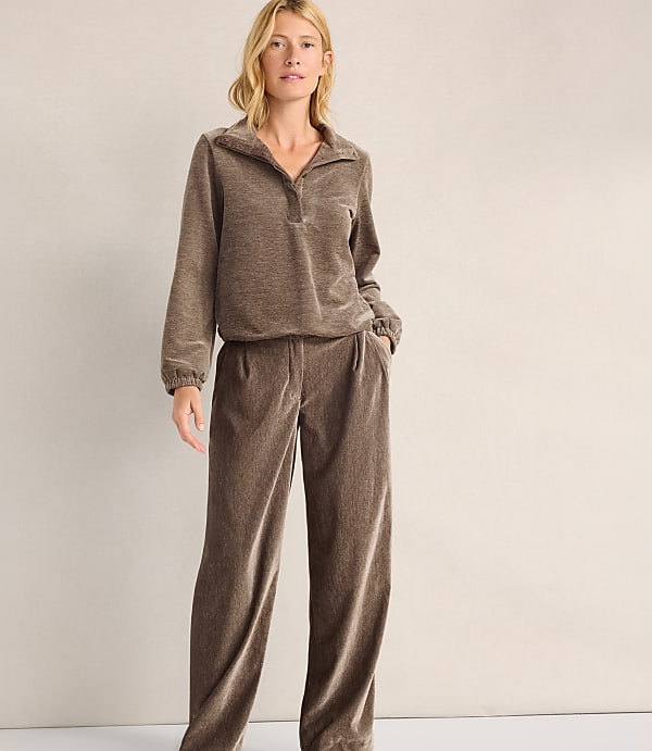 A woman wearing a brown velvet lounge set stands against a plain, light-colored wall.