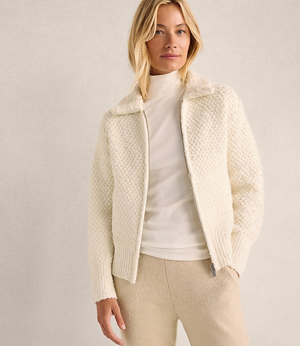 A blonde woman wearing a white textured zip-up jacket and beige trousers stands against a plain, light-colored wall.