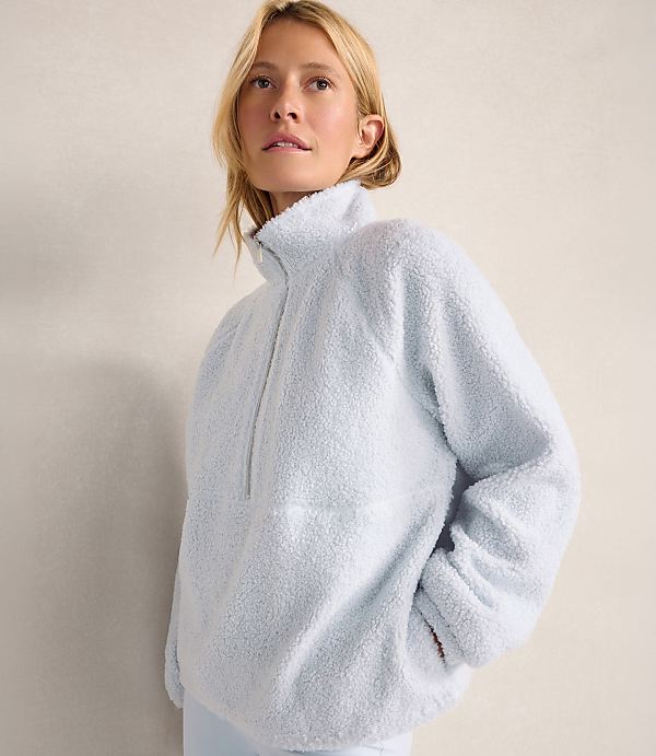 A blonde woman wearing a light blue fuzzy fleece pullover stands against a plain, neutral-toned wall.