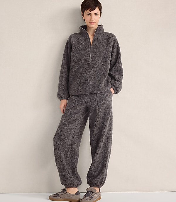 A person wearing a charcoal grey fleece pullover and matching pants stands against a plain, light-colored wall.