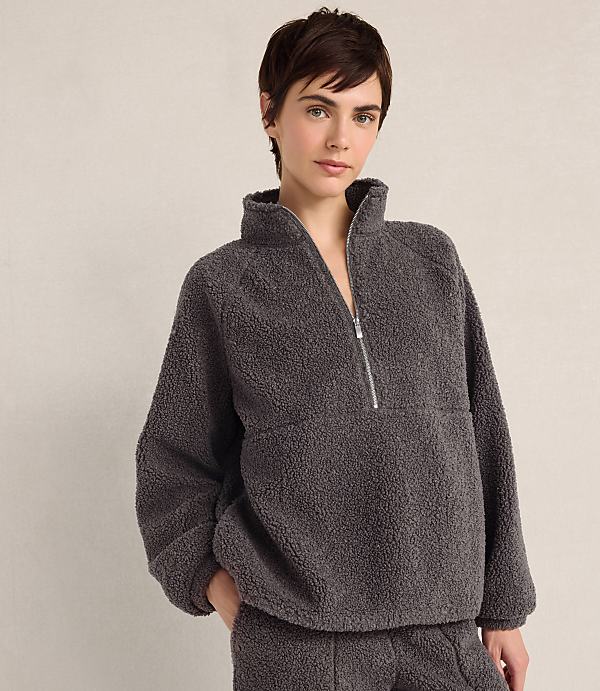 A woman with short brown hair wears a charcoal grey fleece quarter-zip pullover and matching pants against a plain, light-colored background.