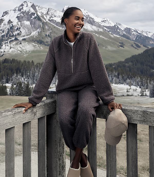 A smiling woman in a charcoal fleece set sits on a wooden railing against a backdrop of snow-capped mountains and evergreen forests.