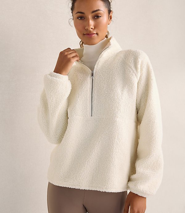 A woman wearing a white fleece pullover and a white turtleneck stands against a plain, neutral-toned background.