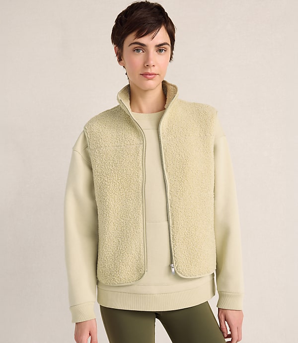A woman with short dark hair wears a cream-colored fleece vest over a matching sweatshirt and olive green leggings against a plain, neutral background.