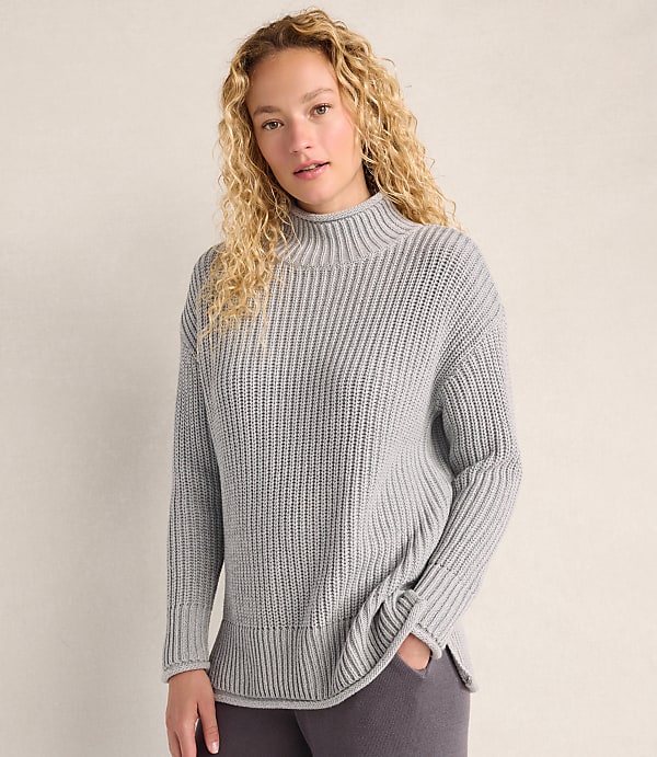 A woman with curly blonde hair wearing a grey ribbed sweater stands against a plain, light-colored wall.