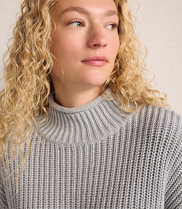A woman with curly blonde hair wearing a grey ribbed sweater stands against a plain, neutral-toned background.
