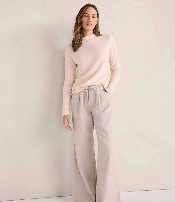 A woman wearing a light pink sweater and wide-leg beige trousers stands against a plain, neutral-colored wall.