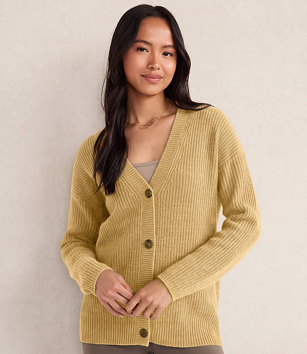 A woman wearing a yellow ribbed cardigan and gold necklaces stands against a plain, light-colored wall.