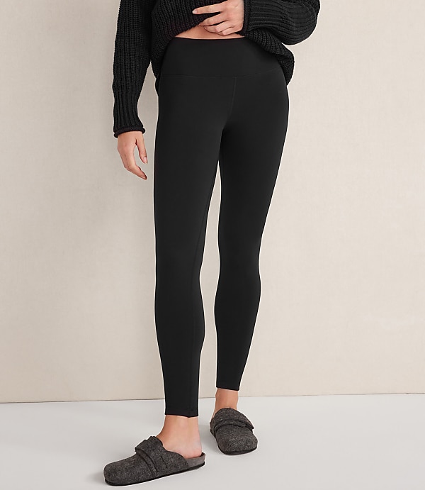 A person wearing a black sweater, black leggings, and grey slippers stands against a plain, light-colored wall.