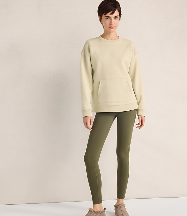 A woman wearing a cream-colored sweatshirt and olive green leggings stands against a plain, neutral-toned wall.