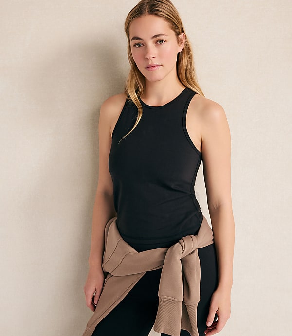 A woman wearing a black tank top and leggings with a tan sweater tied around her waist stands against a plain, light-colored wall.