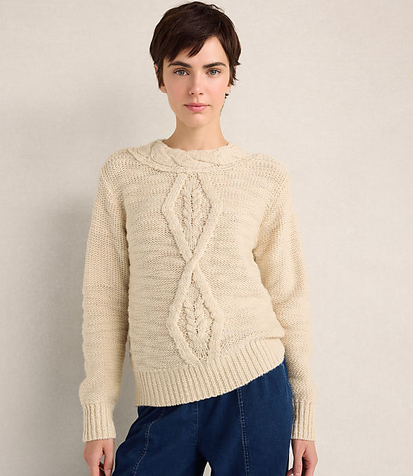 A woman with short brown hair wearing a cream cable-knit sweater and blue pants stands against a plain, neutral-colored wall.