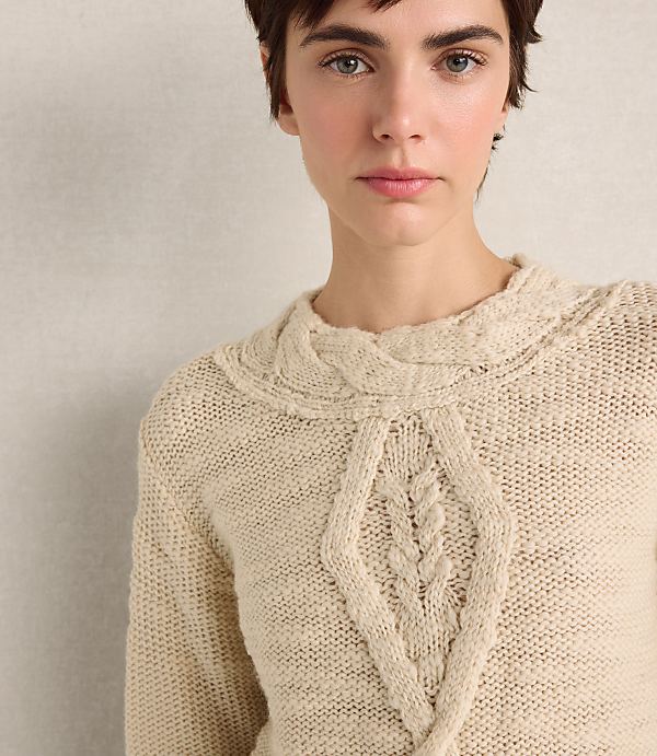 A woman with short dark hair wearing a cream-colored cable-knit sweater stands against a plain, neutral-toned wall.