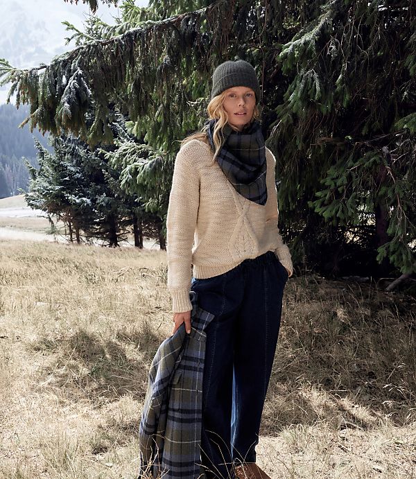 A woman wearing a beanie, cream sweater, and dark trousers stands in a grassy field beneath the branches of evergreen trees.
