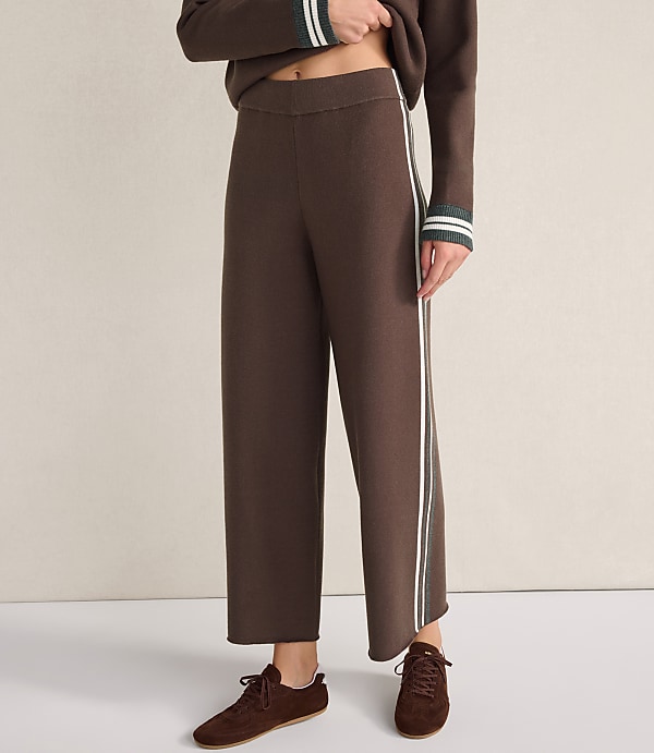 A person wears brown wide-leg trousers with white side stripes and matching brown shoes against a plain, light-colored background.