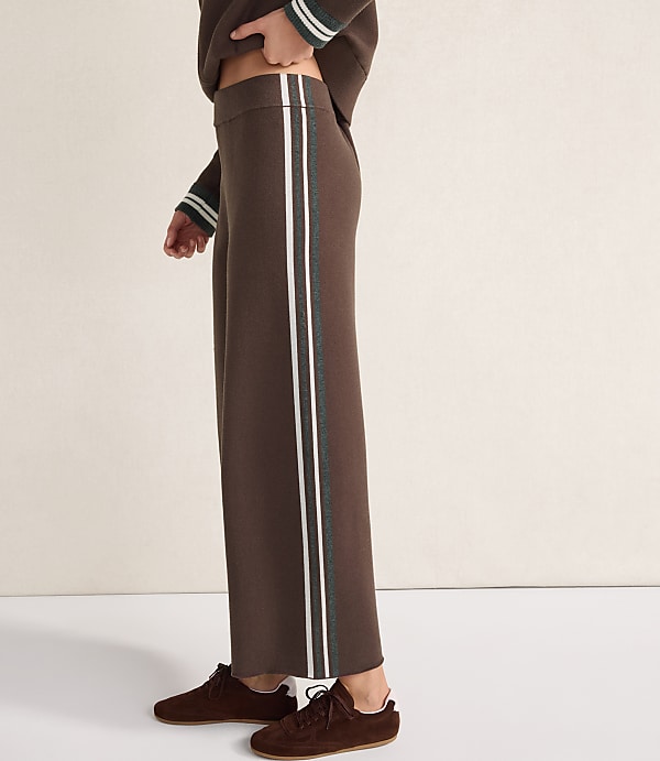 A person wearing brown wide-leg trousers with white and green side stripes and brown suede shoes stands against a plain, light-colored wall.