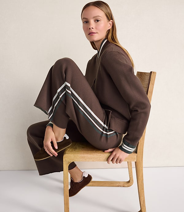 A woman wearing a brown tracksuit and loafers sits on a wooden chair against a plain, light-colored wall.