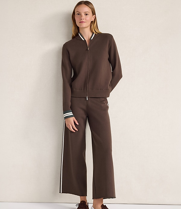 A woman wearing a brown zip-up jacket and matching wide-leg trousers stands against a plain, light-colored wall.