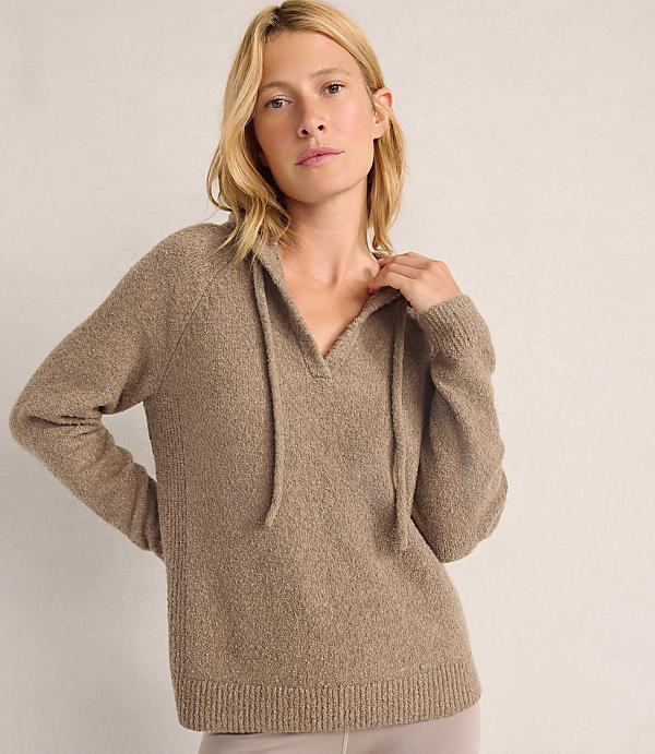 A blonde woman wearing a taupe knit sweater stands against a plain, light-colored background.