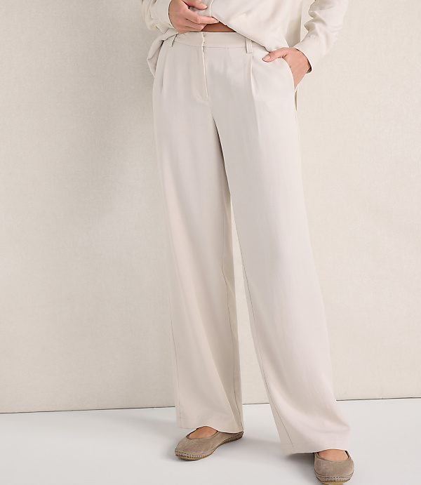 A person wears wide-leg cream trousers and beige flats against a plain, light-colored wall.