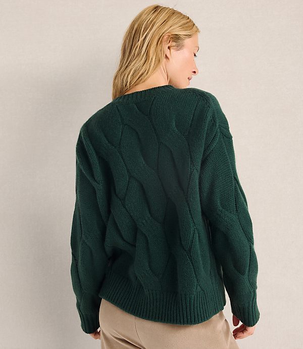 A woman with blonde hair wearing a dark green cable-knit sweater and tan pants stands against a plain, light-colored wall.