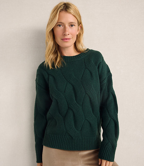 A blonde woman wearing a dark green cable-knit sweater and tan skirt stands against a plain, light-colored wall.