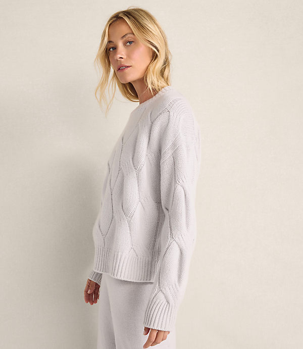 A blonde woman wearing a light-colored cable-knit sweater and matching pants stands against a plain, neutral-toned wall.