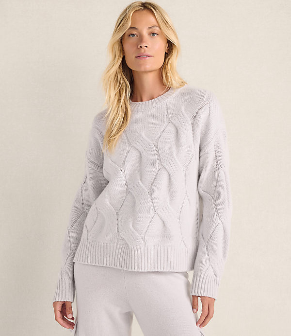 A blonde woman wearing a light purple cable-knit sweater and matching pants stands against a plain, neutral-colored wall.