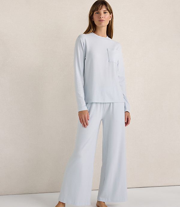 A woman wearing a light blue long-sleeved top and wide-leg pants stands against a plain, neutral-toned wall.