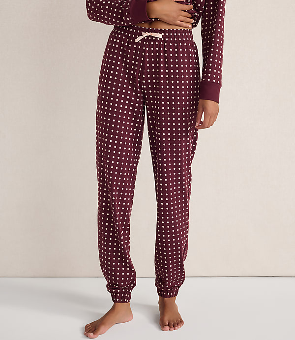 A person wears burgundy polka-dot pajama pants and a matching top against a plain, neutral-colored wall.
