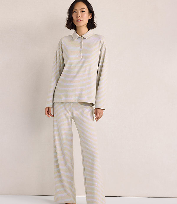 A woman wearing a light-colored, long-sleeved collared shirt and matching wide-leg pants stands against a plain, neutral-toned background.