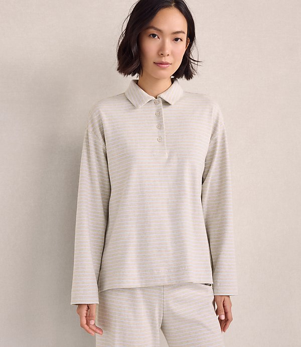 A woman wearing a light-colored striped polo shirt and matching pants stands against a plain, neutral-toned wall.