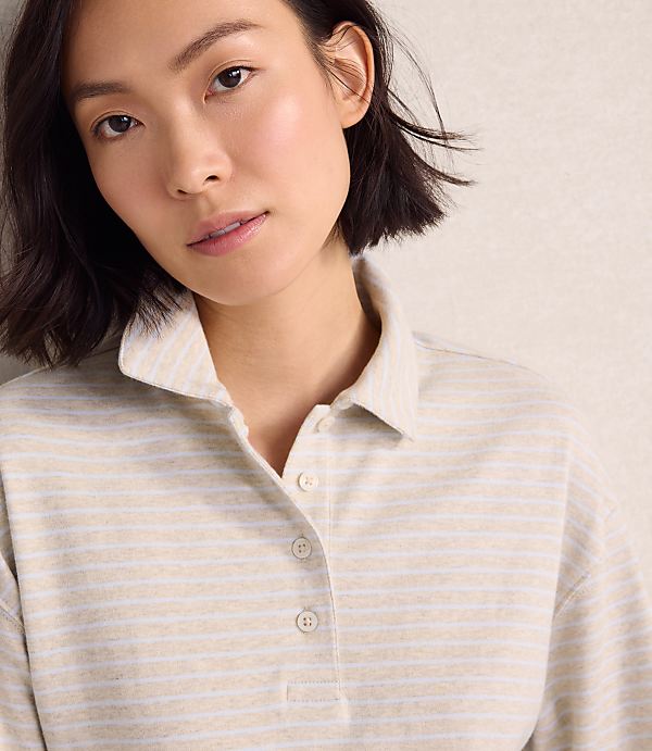 A woman wearing a beige striped polo shirt stands against a plain, light-colored wall.
