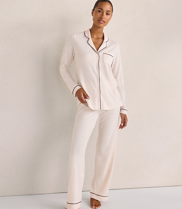 A woman wearing cream-colored pajamas with brown piping stands against a plain, neutral-toned wall.