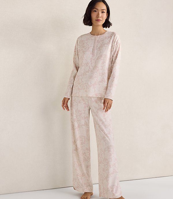 A woman wearing a light pink floral pajama set stands against a plain, neutral-colored wall.