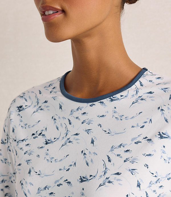 A woman wearing a white floral print shirt stands against a plain, light-colored background.