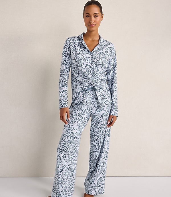 A woman wearing blue and white paisley pajamas stands against a plain, neutral-colored wall.
