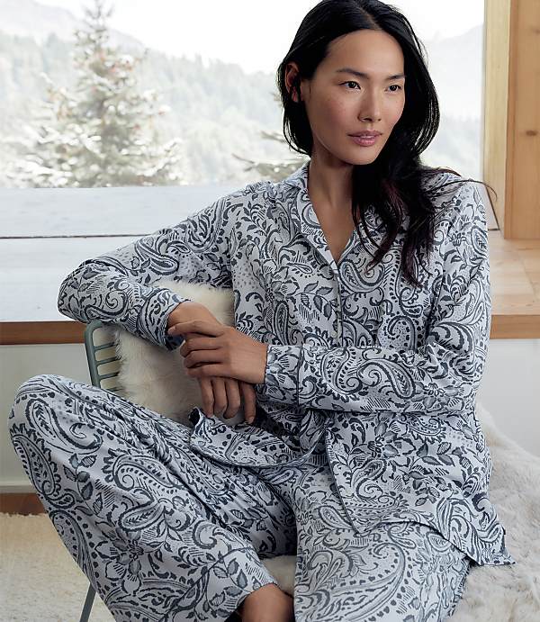 A woman wearing grey and white paisley pajamas sits on a chair in the foreground, with a snowy forest landscape visible through a window in the background.
