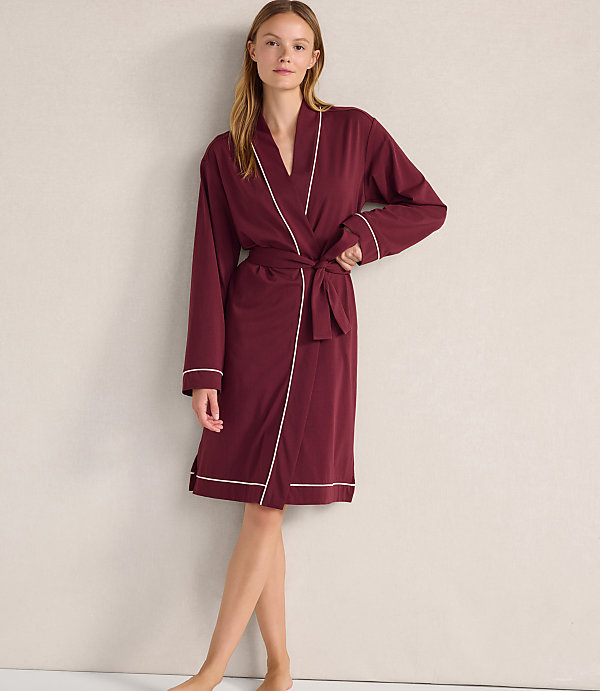 A woman wearing a maroon robe with white trim stands against a plain, light-colored wall.