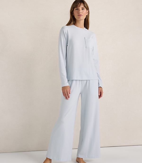 A woman wearing a light blue long-sleeved shirt and matching wide-leg pants stands against a plain, neutral-toned wall.