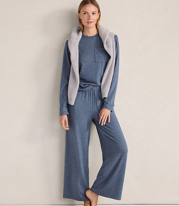 A woman wearing a blue long-sleeved top and wide-leg pants with a grey sweater draped over her shoulders stands against a plain, neutral-colored wall.