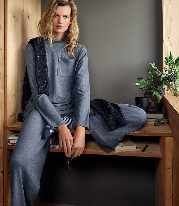 A woman in a grey lounge set sits on a wooden bench against a plain grey wall, with a small potted plant and books nearby.