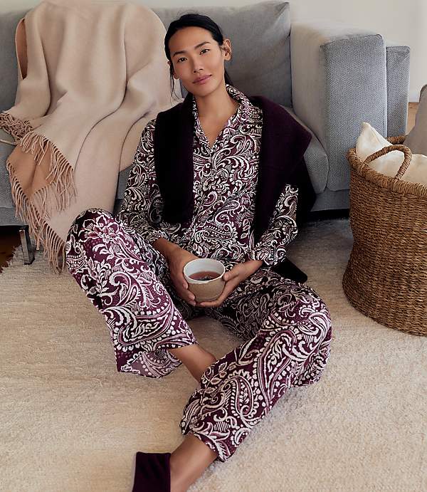 A woman in purple paisley pajamas sits on a white rug in the foreground, with a grey sofa and a woven basket in the background.