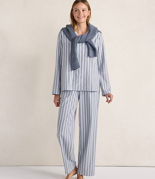 A woman wearing blue and white striped pajamas with a blue sweater draped over her shoulders stands against a plain, light-colored wall.